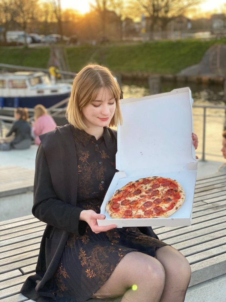 Eine Person hält eine geöffnete Pizzaschachtel mit einer großen Pepperoni-Pizza darin. Sie sitzt entspannt auf einer Bank am Wasser, während ein Boot und andere Menschen im Hintergrund zu sehen sind. Die Szene spielt sich während des Sonnenuntergangs ab, wodurch das warme Licht eine gemütliche Atmosphäre schafft.