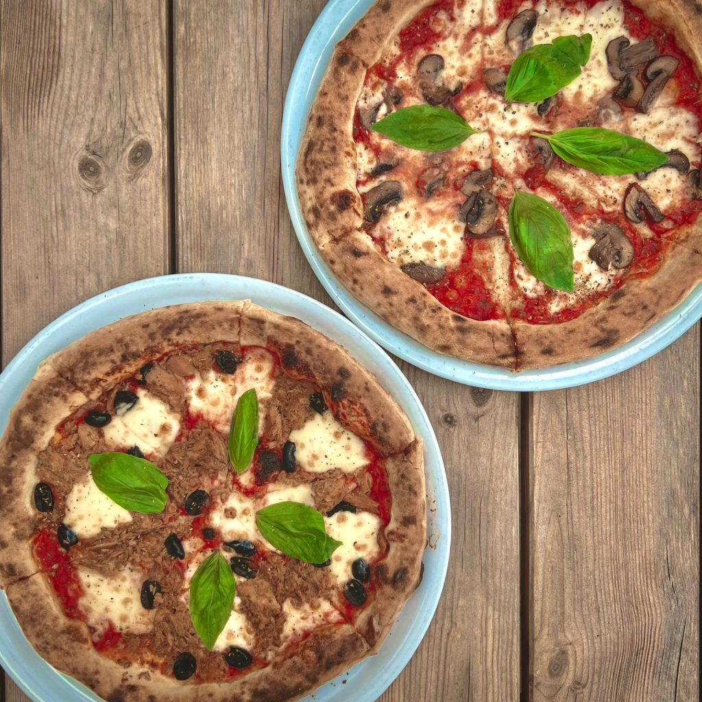 Zwei frisch gebackene Pizzen auf blauen Tellern, serviert auf einer rustikalen Holzoberfläche. Die obere Pizza ist mit Pilzen, Mozzarella, Tomatensoße und frischen Basilikumblättern belegt. Die untere Pizza hat geschreddertes Fleisch, schwarze Oliven, Tomatensoße, Mozzarella und ebenfalls frisches Basilikum als Topping. Der knusprige Teig und die vielfältigen Beläge unterstreichen die kulinarische Vielfalt.